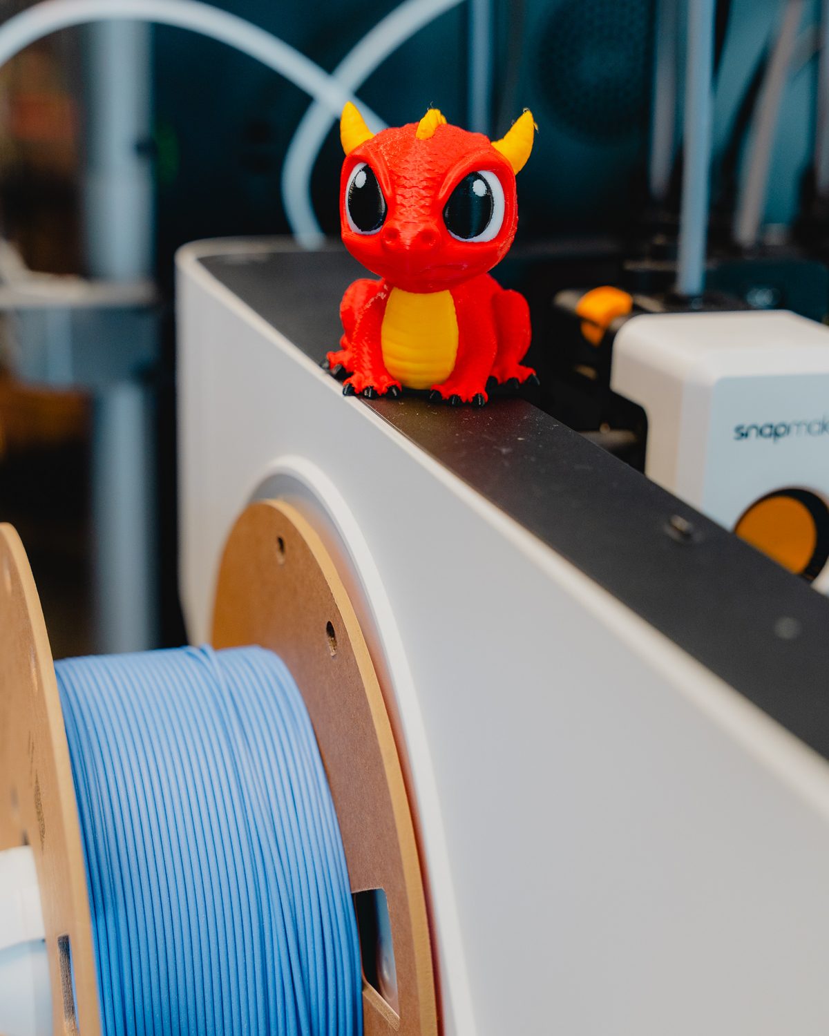 Een rood geprint draakje op een 3D-printer, met een spoel blauw filament ernaast.