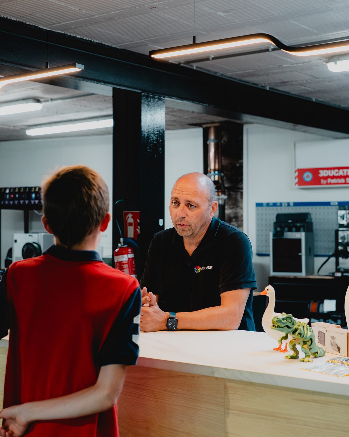 Een medewerker legt aan een jongen uit hoe 3D-printen werkt, aan de toonbank in de winkel.
